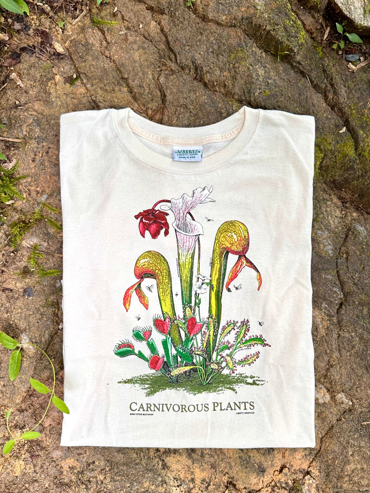 A folded off-white graphic T-shirt laid flat on a textured brown rock outdoors, featuring a colorful illustration of carnivorous plants and the text 'CARNIVOROUS PLANTS' printed below.